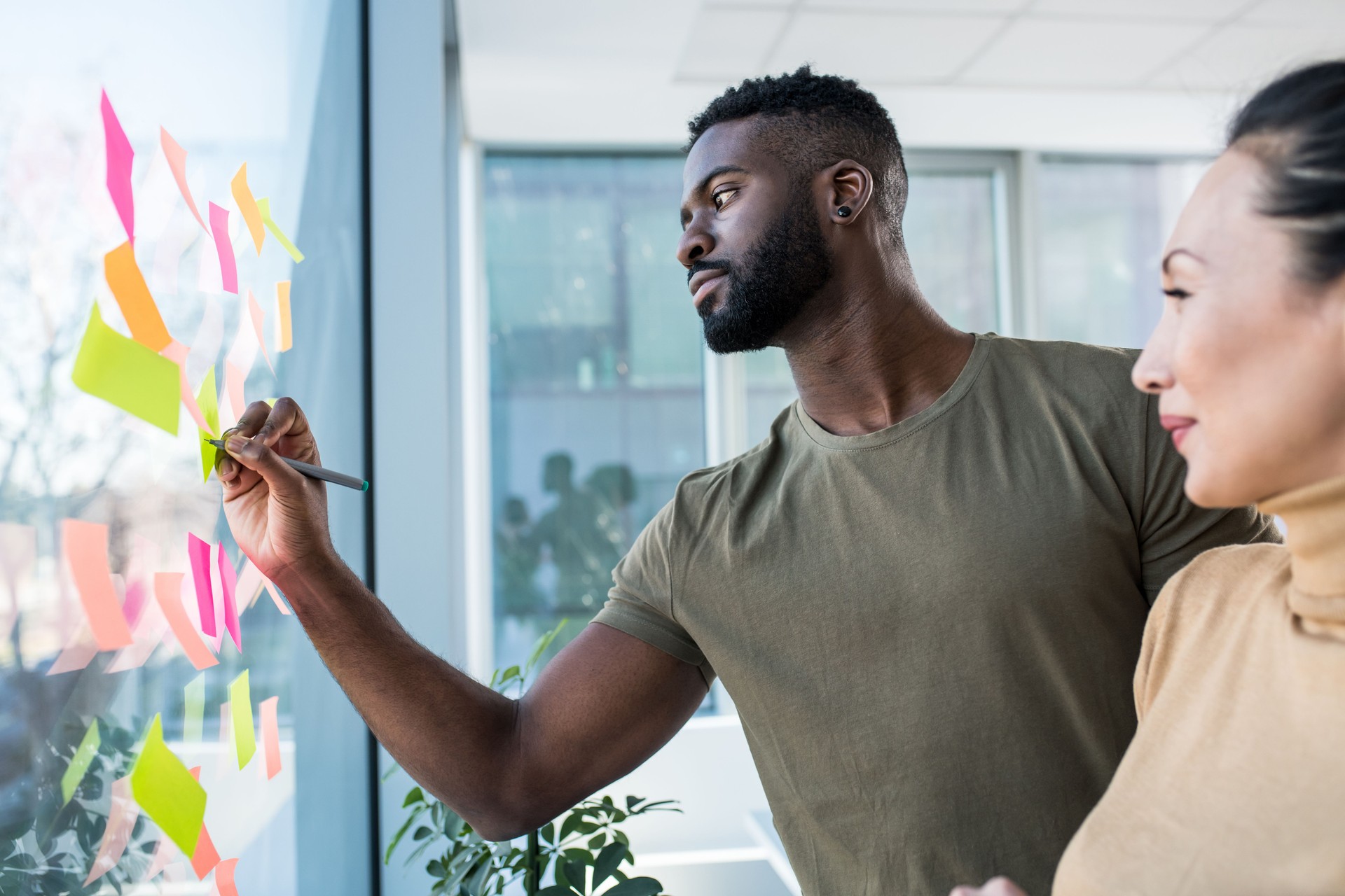 A young African man and an Asian woman brainstorming ideas in the office. There are sticky notes pasted on the large window and they are writing down the ideas.